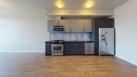 This contemporary single-floor space is designed with clean lines and modern finishes throughout. The open-plan kitchen serves as a central feature, equipped with a full suite of stainless steel appliances including a gas range, built-in microwave, dishwasher, and refrigerator with ice maker. Two-tone cabinetry in light wood and dark charcoal tones pairs with dark countertops and a distinctive vertical mosaic tile backsplash, creating a stylish and functional cooking area. Light hardwood flooring extends across the main living areas, lending warmth and continuity to the space. The full bathroom features a soaking tub with an overhead shower, framed by white subway tiles accented with vertical mosaic tile borders in earth tones, complemented by polished chrome fixtures. A separate half bathroom offers a modern vanity with a dark countertop, framed mirror, and tiled flooring, with a small window allowing natural light to filter in. A dedicated walk-in closet provides practical storage, featuring a full-width hanging rod, overhead shelving, and soft carpeted flooring set against cool light-blue walls. The overall design balances contemporary aesthetics with functional living, offering a cohesive and well-equipped interior.
