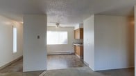 This single-floor space features an open-concept design that connects the living area and kitchen in a functional, straightforward layout. The living area is finished with carpeting and is illuminated by natural light streaming through a large window fitted with vertical blinds. A ceiling fan is centrally positioned overhead, providing both light and air circulation. The textured popcorn ceiling spans throughout, adding a classic finish to the space.

Transitioning from the living area, the kitchen features tile flooring in a warm earth-tone palette, offering a practical and easy-to-maintain surface. Medium-toned wood cabinetry lines the upper walls of the kitchen, providing ample storage space, while a countertop with a built-in cooktop completes the cooking area. A baseboard heater runs along the wall beneath the window, serving as the primary heat source for the space.

The neutral white walls throughout create a clean, bright atmosphere, while the open sightlines between rooms enhance the overall sense of space. The layout is efficient and well-suited for everyday living, with each area clearly defined yet connected within the single-floor footprint.