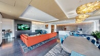 This expansive, single-floor space is a masterclass in contemporary design, seamlessly blending a stylish lounge, dining area, and fully equipped kitchen into one cohesive open-plan layout. The living area anchors the space with a bold terracotta sectional sofa set atop a geometric patterned area rug, complemented by a cobalt blue accent armchair and a sleek coffee table arrangement. A large wall-mounted flat-screen TV is framed by dramatic dark paneling and a striking marble-veined feature wall, adding depth and visual interest. Overhead, sculptural gold pendant lights and layered tray ceilings with warm recessed lighting lend an upscale, curated ambiance throughout. The kitchen is defined by floor-to-ceiling concrete-finish cabinetry with under-cabinet accent lighting, a clean white island with an integrated sink, and stainless steel appliances including a built-in wall oven. A minimalist dining table with teal upholstered chairs bridges the kitchen and lounge zones effortlessly. Large windows and glass doors flood the space with natural light and frame elevated city skyline views, while potted greenery and sheer drapery soften the modern aesthetic. The dark slate-tile flooring ties every element together, creating a polished, inviting atmosphere ideal for both gathering and entertaining.