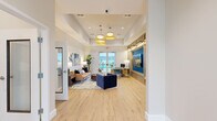 Located at 5901 Myrtle Dr in Wildwood, Florida, this single-floor space serves as a polished and inviting communal area. The entrance greets visitors with light hardwood flooring, recessed lighting, and glass-paneled doors that allow natural light to flow in, leading seamlessly into the main living area.

The living room is the centerpiece of the space, thoughtfully designed with a contemporary coastal aesthetic. A tray ceiling with warm pendant lighting adds architectural detail and a sense of height, while large glass doors offer direct views of an outdoor pool and surrounding landscaping. The room features a built-in wood-paneled accent wall with a bold blue recessed section housing a large flat-screen display, illuminated by sleek picture lights. A built-in shelving unit provides additional storage and display space.

The seating arrangement includes a tan leather sofa and navy blue accent chairs set atop a layered area rug, anchored by a round wood coffee table. Tropical botanical artwork adorns the walls, reinforcing the relaxed Florida ambiance. A workstation with a mid-century-style desk and teal accent chairs is positioned near the pool-facing doors, making the space functional as well as welcoming. Throughout, the light hardwood floors and neutral palette create a bright, airy atmosphere suited for both relaxation and community gatherings.