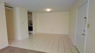 This single-floor space presents a clean, functional layout with a spacious open living area as its centerpiece. The room features light-toned ceramic tile flooring laid in a classic grid pattern, contributing to a bright and airy atmosphere. Neutral cream-colored walls throughout provide a versatile backdrop suitable for a variety of furnishing styles. A flush-mount ceiling light fixture illuminates the main living space evenly.

Adjacent to the living area, a separate kitchen is visible, outfitted with white upper cabinetry and dark-fronted appliances, including what appears to be a range or oven. The kitchen space offers a compact yet practical layout for everyday use. A transition in flooring is noticeable at the entry area, where carpet meets tile, suggesting a defined entryway zone.

The overall ambiance of the space is modest and straightforward, emphasizing clean lines and a neutral palette. The open wall space in the main living area allows for flexible furniture arrangement, making the room adaptable to various needs. The unit's simple design and light color scheme create a sense of openness throughout.