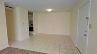 This single-floor space presents a clean, functional layout with a spacious open living area as its centerpiece. The room features light-toned ceramic tile flooring laid in a classic grid pattern, contributing to a bright and airy atmosphere. Neutral cream-colored walls throughout provide a versatile backdrop suitable for a variety of furnishing styles. A flush-mount ceiling light fixture illuminates the main living space evenly.

Adjacent to the living area, a separate kitchen is visible, outfitted with white upper cabinetry and dark-fronted appliances, including what appears to be a range or oven. The kitchen space offers a compact yet practical layout for everyday use. A transition in flooring is noticeable at the entry area, where carpet meets tile, suggesting a defined entryway zone.

The overall ambiance of the space is modest and straightforward, emphasizing clean lines and a neutral palette. The open wall space in the main living area allows for flexible furniture arrangement, making the room adaptable to various needs. The unit's simple design and light color scheme create a sense of openness throughout.