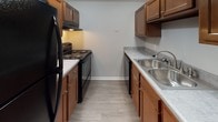 Located at 507 S Main St in Englewood, Ohio, this single-floor residence offers a practical and well-organized layout designed for comfortable everyday living. The galley-style kitchen is efficiently appointed with warm wood cabinetry, light countertops, a stainless steel sink, and black appliances including a refrigerator, electric range, and dishwasher. Light vinyl plank flooring runs throughout the kitchen, providing easy maintenance and a clean aesthetic. The open living area transitions seamlessly from carpet to the kitchen, creating a connected and flexible space. A ceiling fan with integrated lighting adds comfort and function to the main living zone, which also features a closet for additional storage. The bedrooms are carpeted and finished in neutral gray tones, each offering natural light through blinds-covered windows with views of the surrounding greenery. Closets with bifold doors provide dedicated storage in the bedrooms. The consistent use of light gray walls and white trim throughout the unit creates a cohesive, clean look. The overall layout, as seen in the dollhouse view, is well-proportioned with logical room separation, making the space both functional and move-in ready.