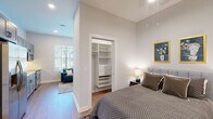 This single-floor unit is designed with a clean, contemporary aesthetic throughout, combining functional layout with modern finishes. The open-concept living and kitchen area serves as the heart of the space, where warm hardwood-style flooring, recessed lighting, and large windows with horizontal blinds create a bright and welcoming atmosphere with views of the surrounding neighborhood.

The kitchen is outfitted with gray shaker-style cabinetry extending to the ceiling, white quartz countertops, a white subway tile backsplash, and a full suite of stainless steel appliances including a refrigerator, electric range, over-the-range microwave, and dishwasher. The living area benefits from notably tall ceilings that lend an airy sense of openness to the space.

The bedroom continues the tall ceiling design and features hardwood-style flooring, recessed lighting, and a built-in closet system with shelving and drawers for ample organized storage. A hallway corridor connects the bedroom to the main living area and also houses an in-unit washer and dryer behind louvered doors.

The bathroom is finished with large-format light gray tile flooring, a dark vanity with white countertop, and a glass-enclosed walk-in shower, maintaining the unit's consistent modern and refined character.