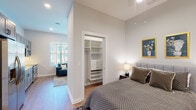 This single-floor unit is designed with a clean, contemporary aesthetic throughout, combining functional layout with modern finishes. The open-concept living and kitchen area serves as the heart of the space, where warm hardwood-style flooring, recessed lighting, and large windows with horizontal blinds create a bright and welcoming atmosphere with views of the surrounding neighborhood.

The kitchen is outfitted with gray shaker-style cabinetry extending to the ceiling, white quartz countertops, a white subway tile backsplash, and a full suite of stainless steel appliances including a refrigerator, electric range, over-the-range microwave, and dishwasher. The living area benefits from notably tall ceilings that lend an airy sense of openness to the space.

The bedroom continues the tall ceiling design and features hardwood-style flooring, recessed lighting, and a built-in closet system with shelving and drawers for ample organized storage. A hallway corridor connects the bedroom to the main living area and also houses an in-unit washer and dryer behind louvered doors.

The bathroom is finished with large-format light gray tile flooring, a dark vanity with white countertop, and a glass-enclosed walk-in shower, maintaining the unit's consistent modern and refined character.