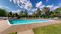 Located at 151 S Bishop Ave in Secane, PA, this outdoor pool area serves as a well-appointed community amenity designed for relaxation and recreation. The centerpiece is a generously sized swimming pool with a curved, freeform shape, featuring classic stainless steel entry railings and steps for easy access. The crystal-clear, blue-tinted water is complemented by a wide concrete and aggregate pool deck that provides ample space for lounging and movement around the perimeter.

The surrounding grounds are thoughtfully landscaped, with neatly trimmed evergreen shrubs, mature deciduous trees, and well-maintained grassy areas that create a lush, park-like atmosphere. A classic white metal fence encloses the entire pool area, providing both safety and a clean aesthetic boundary. Multiple blue lounge chairs and patio umbrellas are arranged along the deck, offering comfortable outdoor seating options.

The backdrop features a multi-story brick residential building with balconies, which frames the space and contributes to the sense of a cohesive community setting. Overhead, open skies allow for abundant natural light throughout the day. The overall ambiance is inviting and well-maintained, making this pool area a standout outdoor feature of the property.