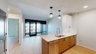 This well-designed one-bedroom, one-bathroom unit offers a thoughtful layout across a single floor, comprising a living room, kitchen, bedroom, bathroom, closet, and hallway. The open-concept living and kitchen area is anchored by a central island with a white quartz countertop, an undermount sink, and warm wood cabinetry that complements the light hardwood flooring found throughout the unit. Stylish globe pendant lights hang above the island, adding a modern accent. The kitchen is equipped with stainless steel appliances, including a full-size refrigerator, microwave, and range, along with a combination of white upper and wood lower cabinetry that maximizes storage. The living area is bright and airy, with floor-to-ceiling sliding glass doors that open to a private balcony, allowing an abundance of natural light and urban views. The bedroom is a comfortable, well-proportioned space featuring a large window with exterior views and clean white walls. The bathroom is finished with dark marble-style tile flooring, a wood vanity with a white countertop, an illuminated mirror, and a glass-enclosed walk-in shower. A dedicated closet is accessible directly from the bathroom, adding practical functionality to the space.
