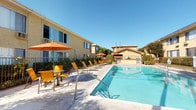 This residential complex offers a thoughtfully designed outdoor environment centered around a large, rectangular swimming pool with clear blue water. The pool area is surrounded by a generous concrete deck furnished with vibrant yellow lounge chairs and shade umbrellas, creating an inviting space for relaxation and recreation. A dedicated BBQ station is conveniently situated nearby, making the outdoor area well-suited for social gatherings. The surrounding buildings feature a classic stucco exterior in warm yellow tones, complemented by solar panels installed on the rooftops — a nod to energy efficiency. Wrought iron fencing encloses the pool area, adding both security and a decorative element to the space. Beyond the pool, the property's grounds are enhanced by lush, well-tended landscaping. Brick-edged garden beds line the walkways, filled with colorful flowering plants including roses, bird of paradise, and other tropical-style foliage, alongside mature palm trees and ornamental shrubs that contribute to a vibrant, garden-like atmosphere. Decorative lamp posts add charm along the pathways. The overall ambiance of the complex is bright, open, and welcoming, with abundant natural light and a warm California aesthetic that emphasizes outdoor living and community amenities.
