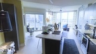 This single-floor residence showcases a thoughtfully designed open-concept layout that integrates the kitchen, dining, and living areas into one cohesive, light-filled space. Floor-to-ceiling windows wrap around the living area, flooding the interior with natural light while framing dramatic urban skyline views, giving the space a distinctly elevated, metropolitan character.

The kitchen serves as a functional centerpiece, anchored by a sleek island with a dark base and light stone countertop, a double undermount sink, and stylish pendant lighting suspended above. The design balances practicality with modern aesthetics, offering bar seating for casual dining.

The dining area sits adjacent to sliding glass doors that open to an outdoor balcony, enhancing the connection between interior and exterior living. The living room beyond is generously proportioned, featuring a ceiling fan and comfortable seating arranged to take full advantage of the panoramic city views through the expansive window walls.

Light hardwood flooring runs throughout, contributing to the airy, contemporary atmosphere. The overall ambiance is modern, luminous, and urban, with an emphasis on open sightlines and natural light.