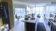 This single-floor residence showcases a thoughtfully designed open-concept layout that integrates the kitchen, dining, and living areas into one cohesive, light-filled space. Floor-to-ceiling windows wrap around the living area, flooding the interior with natural light while framing dramatic urban skyline views, giving the space a distinctly elevated, metropolitan character.

The kitchen serves as a functional centerpiece, anchored by a sleek island with a dark base and light stone countertop, a double undermount sink, and stylish pendant lighting suspended above. The design balances practicality with modern aesthetics, offering bar seating for casual dining.

The dining area sits adjacent to sliding glass doors that open to an outdoor balcony, enhancing the connection between interior and exterior living. The living room beyond is generously proportioned, featuring a ceiling fan and comfortable seating arranged to take full advantage of the panoramic city views through the expansive window walls.

Light hardwood flooring runs throughout, contributing to the airy, contemporary atmosphere. The overall ambiance is modern, luminous, and urban, with an emphasis on open sightlines and natural light.