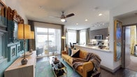 This single-floor space showcases a thoughtfully designed open-concept layout that seamlessly connects the living area and kitchen into one cohesive, functional environment. The living area serves as the centerpiece, featuring rich hardwood-style flooring, a vibrant teal area rug, and a bold mustard-yellow sofa accented with decorative pillows and a cozy throw blanket. A sleek glass-and-gold coffee table adds a refined touch, while a ceiling fan with an integrated light fixture overhead provides both comfort and ambiance. Recessed lighting and track lighting throughout ensure the space is well-illuminated at all times.

Large sliding glass doors flood the living area with abundant natural light and open directly onto a private balcony, where additional seating is visible. Elegant draping curtains frame the openings, softening the transition between interior and exterior.

The kitchen, positioned adjacent to the living space, features modern gray cabinetry, light stone countertops with a breakfast bar overhang, stainless steel appliances including a microwave and range, and under-cabinet task lighting. The open pass-through design keeps the kitchen visually connected to the living area, enhancing the overall sense of openness and flow throughout the space.