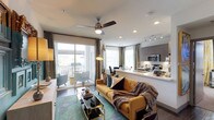 This single-floor space showcases a thoughtfully designed open-concept layout that seamlessly connects the living area and kitchen into one cohesive, functional environment. The living area serves as the centerpiece, featuring rich hardwood-style flooring, a vibrant teal area rug, and a bold mustard-yellow sofa accented with decorative pillows and a cozy throw blanket. A sleek glass-and-gold coffee table adds a refined touch, while a ceiling fan with an integrated light fixture overhead provides both comfort and ambiance. Recessed lighting and track lighting throughout ensure the space is well-illuminated at all times.

Large sliding glass doors flood the living area with abundant natural light and open directly onto a private balcony, where additional seating is visible. Elegant draping curtains frame the openings, softening the transition between interior and exterior.

The kitchen, positioned adjacent to the living space, features modern gray cabinetry, light stone countertops with a breakfast bar overhang, stainless steel appliances including a microwave and range, and under-cabinet task lighting. The open pass-through design keeps the kitchen visually connected to the living area, enhancing the overall sense of openness and flow throughout the space.