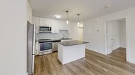 Located at 4981 Bathurst Street in Toronto's North York neighbourhood, this one-bedroom unit offers a thoughtfully designed layout with modern finishes throughout. Light-toned hardwood-style flooring runs continuously across the entire space, creating a cohesive and clean aesthetic.

The kitchen is a standout feature, showcasing white shaker-style cabinetry extending to the ceiling for ample storage, paired with a light subway tile backsplash and dark countertops. A central island with a dark countertop surface provides additional workspace and casual dining potential. Stainless steel appliances — including a French door refrigerator, electric range, and over-the-range microwave — complete the modern, functional kitchen. Dual pendant lights hang above the island, adding a stylish finishing touch.

The bedroom is a comfortable, well-proportioned room finished in crisp white with clean baseboards. Two windows allow natural light to filter in and offer views of greenery outside, creating a calm and pleasant atmosphere. A wall-mounted heating unit ensures year-round comfort.

The overall design of the unit is bright, clean, and contemporary, with a neutral palette that lends itself to a variety of interior styles. The building itself is a multi-storey residential structure set in a mature, tree-lined setting in Toronto.
