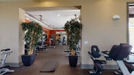 This single-level fitness center is organized into several distinct workout zones, offering a comprehensive range of exercise options. The space features a bold design aesthetic, with deep orange and red accent walls contrasting against neutral beige tones throughout. Rubber flooring provides a durable, safety-conscious surface across all areas.

The cardio zone is positioned near large, grid-paned windows that allow generous natural light and outdoor views of surrounding landscaping and palm trees. A tray ceiling with a ceiling fan adds architectural interest to this section. Cardio equipment includes treadmills, elliptical trainers, stationary bikes, and spin cycles.

The strength training area features a full power rack with a wall-mounted flat-screen TV, a weight bench, and an assortment of plate weights. Floor-to-ceiling mirrors visually expand the space. A separate section houses multiple selectorized cable machines and resistance equipment positioned in front of additional large windows.

Tall decorative bamboo plants are strategically placed throughout, adding a natural, polished element to the environment. Recessed lighting and ceiling fans ensure comfort across all zones, while the overall layout supports an efficient, well-rounded training experience.
