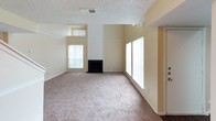 This two-story residence offers a practical and straightforward layout across both levels. The main living area is a standout feature, boasting vaulted ceilings that rise to impressive heights, flanked by multiple windows that allow generous natural light to fill the space. A black fireplace serves as the room's focal point, adding a functional and aesthetic centerpiece to the open, carpeted living area.

The kitchen is equipped with black appliances, including a coil-burner range with an overhead exhaust hood, dark cabinetry, and speckled countertops, complemented by tile flooring. A staircase with a white railing connects the main level to the upper floor, where a carpeted loft-style landing area overlooks the lower level, providing additional flexible living space.

The bedrooms are carpeted and finished in neutral tones, each featuring built-in closets with shelving for organized storage. One bedroom includes a walk-in closet with multiple shelving tiers. The bathroom features a full tub-shower combination with white tile surround, chrome fixtures, and dark wood-look flooring. Textured ceilings are consistent throughout, contributing to the home's cohesive and understated aesthetic.