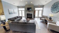 This single-floor communal lounge is thoughtfully designed to foster comfort and social interaction. The space centers around a classic white-mantled fireplace set against a gray accent wall, above which a wall-mounted flat-screen television is positioned for easy viewing. Vaulted ceilings give the room a spacious, open feel, while recessed lighting supplements the generous natural light flowing in through multiple large, grid-paned windows and glass-paneled French doors flanking the fireplace on both sides.

The flooring consists of rich, dark hardwood planks that contrast elegantly with the soft gray and white color palette of the walls and trim. The seating arrangement includes a large sectional sofa and additional accent chairs, all in coordinating gray tones, centered around a round wooden coffee table and anchored by a patterned area rug. Decorative elements such as dried botanicals and minimalist artwork contribute to the room's contemporary yet inviting atmosphere. The overall design balances modern aesthetics with functional comfort, making it well-suited as a shared gathering space.