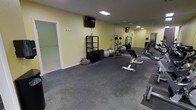 This dedicated fitness space is thoughtfully designed to support a wide range of exercise routines within a single-floor layout. The room features durable rubber flooring, ideal for high-impact workouts and heavy equipment use, providing both safety and stability underfoot.

The space is outfitted with a variety of workout equipment, including a weight bench, free weights, exercise balls, and foam rollers, catering to both strength training and flexibility work. A multi-shelf storage rack keeps accessories neatly organized, while a wall-mounted television adds entertainment or instructional viewing during sessions.

A floor-to-ceiling mirror spans one wall, visually expanding the room and allowing users to monitor their form during exercises. Recessed and overhead lighting illuminate the space evenly, creating a bright and energizing atmosphere. The soft yellow walls contribute a clean, motivating aesthetic throughout.

An adjoining room, accessible through a doorway, appears to offer additional open floor space — potentially suited for stretching, yoga, or mat-based workouts. The overall layout is practical and efficient, maximizing usable floor area while keeping the environment uncluttered and focused on fitness functionality.