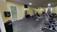 This dedicated fitness space is thoughtfully designed to support a wide range of exercise routines within a single-floor layout. The room features durable rubber flooring, ideal for high-impact workouts and heavy equipment use, providing both safety and stability underfoot.

The space is outfitted with a variety of workout equipment, including a weight bench, free weights, exercise balls, and foam rollers, catering to both strength training and flexibility work. A multi-shelf storage rack keeps accessories neatly organized, while a wall-mounted television adds entertainment or instructional viewing during sessions.

A floor-to-ceiling mirror spans one wall, visually expanding the room and allowing users to monitor their form during exercises. Recessed and overhead lighting illuminate the space evenly, creating a bright and energizing atmosphere. The soft yellow walls contribute a clean, motivating aesthetic throughout.

An adjoining room, accessible through a doorway, appears to offer additional open floor space — potentially suited for stretching, yoga, or mat-based workouts. The overall layout is practical and efficient, maximizing usable floor area while keeping the environment uncluttered and focused on fitness functionality.