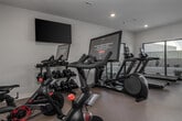 This fitness center is designed to support a wide range of workout routines within a spacious, single-floor layout. The room is bathed in natural light courtesy of large sliding glass windows and picture windows that line multiple walls, offering outdoor views and an open, airy ambiance. Recessed lighting throughout the ceiling ensures consistent illumination across the entire floor.

The cardio section is generously equipped with multiple treadmills featuring integrated touchscreen displays, elliptical trainers, and stationary cycling bikes with individual monitor screens for interactive workouts. A wall-mounted flat-screen television provides additional entertainment options for cardio sessions.

Strength and functional training are well-represented with a multi-station cable machine, a dedicated functional training rig stocked with kettlebells, medicine balls, resistance bands, TRX suspension trainers, and battle ropes. A tiered rack holds a full set of dumbbells, while stability balls and exercise balls are neatly organized on a wall-mounted storage rack.

A weight bench and mat area provide space for stretching and floor exercises. Mirrors are strategically placed to assist with form and technique. The flooring throughout is a durable, neutral-toned surface suited for high-traffic fitness use. The overall design is clean, modern, and functional, making this fitness center a comprehensive facility for diverse training needs.