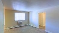 This single-floor residential unit offers a clean, functional layout designed for comfortable everyday living. The main living area is finished with wall-to-wall carpeting that adds warmth underfoot, while neutral-toned walls create a bright and versatile backdrop throughout the space. A window fitted with vertical blinds allows natural light to filter in while providing privacy, contributing to the room's airy feel. Climate control is well-addressed with a wall-mounted air conditioning unit and baseboard heating running along the perimeter of the room, ensuring year-round comfort regardless of the season. The interior doorway visible from the main room leads to additional areas of the unit, suggesting a practical and connected floor plan. Smooth ceilings and clean white trim work frame each room with a simple, tidy finish. The overall ambiance is understated and move-in ready, offering a neutral canvas well-suited for a variety of furnishing styles. The space is straightforward in its design, prioritizing functionality and ease of living throughout its single-level layout.