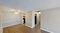 This single-story unit is designed with a practical, open layout that connects the main living areas in a straightforward and functional manner. Throughout the common areas, wood-look plank flooring in warm, medium-toned hues provides a cohesive foundation, complemented by neutral gray-beige walls and crisp white baseboards. Textured ceilings are consistent throughout the space.

The living area is generously proportioned, featuring a ceiling fan with a light fixture and a sliding glass door fitted with vertical blinds, allowing natural light to filter in while providing access to the outdoors. The space flows directly toward the kitchen, which is partially separated by a half-wall partition. The kitchen is equipped with black appliances, including a range and dishwasher, paired with white cabinetry and light countertops, all set on matching wood-look flooring.

The bedrooms feature soft, neutral carpet and are well-sized with clean walls and textured ceilings. One bedroom includes a window with blinds, admitting natural light. The bedroom area connects to a hallway with closet storage, and a bathroom area is accessible nearby, featuring a vanity with a light countertop and additional storage closets. The overall ambiance is clean, neutral, and move-in ready.