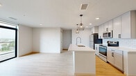 This single-floor unit showcases a clean, contemporary design with light wood-style flooring and crisp white walls that run throughout the entire space. The open-concept kitchen and living area serves as the heart of the unit, anchored by a large white quartz island with an undermount sink and a flexible spray faucet. Geometric wire pendant lights hang above the island, adding an industrial-modern accent. The kitchen is well-appointed with light gray shaker-style cabinetry extending to the ceiling, a white subway tile backsplash, and a full suite of stainless steel appliances including a refrigerator, electric range, and over-the-range microwave. Floor-to-ceiling sliding glass doors in the living area open directly onto a wood-deck balcony, offering outdoor views and flooding the space with natural light. Additional rooms include multiple bedrooms, each benefiting from large black-framed windows with elevated exterior views. A hallway connects the bedrooms to a full bathroom featuring a white vanity with quartz countertop, wood-look flooring, matte black hardware, and a tub-shower combination. A walk-in closet provides ample storage, completing the thoughtfully designed layout.
