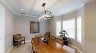 This single-story space showcases a well-appointed dining area that combines classic and contemporary design elements. The room is anchored by a substantial rectangular wood dining table surrounded by upholstered chairs, offering generous seating capacity for gatherings. Above, a striking geometric pendant light fixture with an open metal frame and exposed bulbs serves as a focal point, drawing the eye upward toward the crown-molded ceiling finished in crisp white. The neutral gray walls provide a versatile backdrop, complemented by clean white baseboards that frame the light-toned flooring throughout. Natural light filters into the space through white plantation-shuttered windows, creating a bright and welcoming atmosphere. A large, textured abstract artwork adds visual depth and a touch of warmth to the wall, while a tall dark vase with lush greenery introduces a natural element near the window. A telephone on the table suggests the room may also function as a meeting or conference area. Overall, the space balances refined elegance with practical comfort, making it well-suited for both formal dining and professional gatherings.