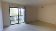 This single-floor space features a carpeted living area with neutral-toned walls and a clean, understated design. The room is brightened by a large sliding glass door that fills the space with natural light while providing direct access to a private balcony. The balcony is enclosed by a dark railing and overlooks a pleasant outdoor setting featuring mature trees and green landscaping, creating a serene and inviting atmosphere. The wall-to-wall carpeting in a light beige tone adds warmth underfoot, complementing the neutral palette of the walls and ceiling. A ceiling vent is visible, indicating a central HVAC system for year-round climate control. The open layout of the living area offers flexible arrangement possibilities, making it adaptable to a variety of furniture configurations. Vertical blinds frame the sliding door, providing privacy and light control as needed. Overall, the space presents a functional and comfortable environment with an emphasis on natural light and outdoor connectivity.