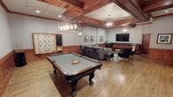 This single-floor entertainment lounge is thoughtfully designed to offer a refined yet relaxed social atmosphere. The centerpiece of the space is a full-size billiards table with ornate carved legs and a slate-gray felt surface, positioned beneath a pair of sophisticated lantern-style pendant chandeliers that cast warm, inviting light throughout the room. The ceiling features rich, dark wood coffered beams that add architectural depth and character, complementing the warm hardwood flooring below. Along one wall, a large flat-screen television is mounted above a built-in credenza with shelving and cabinetry, flanked by a comfortable lounge seating arrangement with gray upholstered sofas and accent chairs — ideal for gathering and conversation. A counter-height bar area with bar stools provides additional seating nearby. On the opposite wall, a large decorative Scrabble board serves as a striking and playful art piece, reinforcing the game-room theme. Subtle wall sconces and framed artwork contribute to the polished, club-like ambiance. The overall design balances casual recreation with tasteful, upscale finishes, making this space well-suited for both lively social events and relaxed leisure.