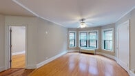 This single-floor unit showcases classic architectural details and a cohesive design throughout. Warm hardwood floors extend across every room, complemented by crisp white crown molding and baseboards that add a polished, traditional character to the space.

The spacious living area is a standout feature, anchored by a curved bay window arrangement that fills the room with natural light and offers views of the surrounding urban streetscape. A ceiling fan with integrated lighting and a cast-iron radiator beneath the windows complete this inviting room.

The layout flows naturally into additional rooms, including two well-proportioned bedrooms, each outfitted with ceiling fans and generous windows that bring in natural light. The rooms share the same warm hardwood flooring and neutral gray-toned walls found throughout the unit.

The kitchen features light maple cabinetry, granite countertops with subtle speckled patterning, a stainless steel sink, and stainless steel appliances including a dishwasher and refrigerator, offering both function and style.

The bathroom is clean and bright, finished with light tile flooring, white subway tile surrounding the full soaking tub, a vanity with mirror, and a window that provides natural ventilation. The overall ambiance of the space is bright, airy, and move-in ready.