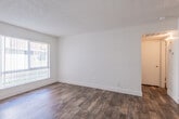 This single-floor unit presents a straightforward, functional layout with a clean and neutral aesthetic throughout. The main living area is generously proportioned, featuring warm wood-look flooring that extends across much of the space. White walls and a textured popcorn ceiling create a bright, open feel, while large windows fitted with vertical blinds allow ample natural light to filter in. A ceiling fan is installed in the living area, adding a practical touch to the space.

The kitchen is efficiently arranged in a galley-style configuration, with white upper and lower cabinetry providing substantial storage. Speckled laminate countertops run along both sides, complemented by a stainless steel electric range and a single-basin sink. Wood-look flooring continues seamlessly from the living area into the kitchen, maintaining a cohesive look throughout.

The bedroom is carpeted in a neutral gray tone, offering a softer underfoot feel, and is served by a window with vertical blinds that provides a view of the surrounding greenery. The full bathroom is compact and functional, featuring a combined tub and shower, toilet, and hardwood-tone flooring. A programmable thermostat is conveniently located in the hallway, adding a modern, energy-efficient element to the unit.