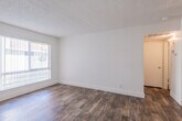 This single-floor unit presents a straightforward, functional layout with a clean and neutral aesthetic throughout. The main living area is generously proportioned, featuring warm wood-look flooring that extends across much of the space. White walls and a textured popcorn ceiling create a bright, open feel, while large windows fitted with vertical blinds allow ample natural light to filter in. A ceiling fan is installed in the living area, adding a practical touch to the space.

The kitchen is efficiently arranged in a galley-style configuration, with white upper and lower cabinetry providing substantial storage. Speckled laminate countertops run along both sides, complemented by a stainless steel electric range and a single-basin sink. Wood-look flooring continues seamlessly from the living area into the kitchen, maintaining a cohesive look throughout.

The bedroom is carpeted in a neutral gray tone, offering a softer underfoot feel, and is served by a window with vertical blinds that provides a view of the surrounding greenery. The full bathroom is compact and functional, featuring a combined tub and shower, toilet, and hardwood-tone flooring. A programmable thermostat is conveniently located in the hallway, adding a modern, energy-efficient element to the unit.