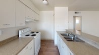 This single-floor residence offers a clean and practical layout designed for comfortable everyday living. The kitchen is a standout feature, designed in a classic galley configuration that maximizes efficiency and storage. White cabinetry lines both sides of the kitchen, providing generous storage space above and below, while speckled laminate countertops run the full length of both walls. On one side, a white coil-burner electric range sits beneath an overhead exhaust hood and upper cabinets, while the opposite side features a double-basin stainless steel sink with a standard faucet. Warm-toned wood-look flooring runs throughout, adding a sense of continuity and warmth to the space. Overhead, a flush-mount ceiling light provides even illumination. A visible HVAC vent in the ceiling suggests a central climate control system. At the far end of the kitchen, an opening leads into an adjacent room where natural light filters in through a window, hinting at a bright and connected living area beyond. The overall aesthetic is neutral and well-maintained, offering a versatile backdrop that is easy to personalize.