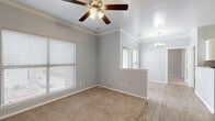 This thoughtfully designed single-floor unit offers a comfortable and functional layout across its one-bedroom, one-bathroom floor plan. The living area features soft carpeting, crown molding, and a ceiling fan with integrated lighting, while multiple windows with blinds allow for ample natural light. A half-wall partition separates the living space from the kitchen, which is outfitted with crisp white shaker-style cabinetry, a stone tile backsplash, dark countertops, and black appliances including a range, microwave, and refrigerator. Wood-look flooring runs throughout the kitchen and entryway, providing a clean contrast to the carpeted areas. The bedroom is a well-sized, light-filled room with a ceiling fan, plush carpeting, and multiple windows offering outdoor views. It connects to a dedicated walk-in closet equipped with multi-tiered wire shelving for organized storage. The bathroom features wood-look flooring, a single vanity with a mirror, and a full bathtub with a shower. Neutral gray walls throughout the unit create a calm, cohesive aesthetic that is both modern and inviting.