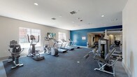 This fitness center is thoughtfully designed to support a full range of workout routines across multiple interconnected zones. The main cardio area features a row of treadmills, ellipticals, and stair climbers arranged beneath recessed lighting and wall-mounted flat-screen TVs, with large windows providing abundant natural light and outdoor views. A bold blue accent wall adds a dynamic visual element to this energetic space. The flooring throughout is covered in durable, dark gray carpet suited for high-traffic exercise use. An adjoining strength training area houses a multi-station cable machine, a leg press machine, weight benches, and a full rack of dumbbells, offering a comprehensive selection of resistance equipment. A dedicated flexibility and functional training zone includes stability balls of varying sizes displayed on a wall-mounted rack, a flat bench, resistance bands, and a large wall mirror accompanied by a mounted TV for guided workout viewing. The space benefits from recessed ceiling lighting throughout, neutral gray and white walls, and clearly defined zones that allow multiple users to exercise simultaneously without interference. Multiple entry and exit points provide convenient access throughout the facility.