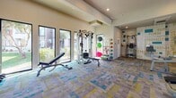 This modern fitness center is thoughtfully designed to support a full range of workout routines within a single, open-plan floor. The space is brightened by large windows that allow ample natural light to pour in, offering views of the surrounding outdoor greenery and creating an energizing atmosphere for exercise.

The colorful, patterned carpet tiles in shades of teal, yellow, and gray add a lively visual character to the floor throughout the space. A striking feature wall is adorned with bold motivational typography — words like "Wellness," "Strength," "Endurance," and "Form" — rendered in teal and yellow tones that complement the overall design aesthetic.

The fitness center is well-stocked with a comprehensive selection of equipment. The cardio zone includes multiple treadmills, elliptical machines, and a recumbent bike, each positioned near windows for an inspiring workout environment. The strength training area offers a cable/functional trainer, a weight bench with free weights, and a multi-station cable machine. A robust free weights section features a full rack of dumbbells and an extensive collection of kettlebells in varying sizes. Stability and medicine balls round out the functional fitness offerings.

Large mirrors are strategically placed to enhance the sense of openness and allow users to monitor their form. Recessed lighting and wall-mounted televisions further elevate the experience, making this fitness center a comprehensive and inviting destination for all fitness levels.