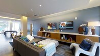 This single-story space presents a contemporary lounge or living area designed with both comfort and functionality in mind. The room is finished with smooth, cool-toned grey walls that lend a calm, modern ambiance throughout. Recessed lighting is embedded in the ceiling, complemented by an elegant tray ceiling detail that adds architectural depth and subtle visual interest overhead.

A standout feature is the wall-mounted entertainment setup, which includes two flat-screen TVs positioned side by side, catering to a multi-screen viewing experience. Beneath the screens, a long, low-profile built-in media console constructed from warm wood tones provides ample open shelving and closed cabinetry. The shelving is styled with decorative vases, books, and accent pieces, adding personality to the space.

Seating consists of a generously sized sectional sofa in a neutral grey upholstery, dressed with colorful accent pillows in teal, green, and yellow. A tufted white ottoman serves as a central coffee table and doubles as additional seating. A patterned area rug anchors the seating arrangement, and a doorway in the background suggests additional rooms beyond, offering connectivity throughout the layout.