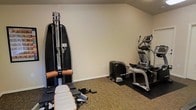 This single-floor fitness space is purpose-built for personal training and exercise, offering a well-organized layout that accommodates a variety of workout styles. The room features neutral-toned walls and low-profile carpeted flooring complemented by protective rubber floor mats beneath the equipment, providing both comfort and stability during use. A sloped ceiling adds architectural character while maintaining an open and functional atmosphere. The space is outfitted with professional-grade fitness equipment, including a SportsArt multi-function cable and strength training machine, ideal for a wide range of resistance exercises. An elliptical cross-trainer offers low-impact cardiovascular conditioning, while an additional compact cardio unit rounds out the equipment lineup. The machines are arranged to allow clear movement between stations, making the room efficient and user-friendly. Neutral wall colors and clean lines keep the environment focused and distraction-free, encouraging productivity during workouts. This space is well-suited for individuals seeking a private, fully equipped fitness area with the convenience of professional equipment in a dedicated room.