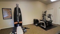 This single-floor fitness space is purpose-built for personal training and exercise, offering a well-organized layout that accommodates a variety of workout styles. The room features neutral-toned walls and low-profile carpeted flooring complemented by protective rubber floor mats beneath the equipment, providing both comfort and stability during use. A sloped ceiling adds architectural character while maintaining an open and functional atmosphere. The space is outfitted with professional-grade fitness equipment, including a SportsArt multi-function cable and strength training machine, ideal for a wide range of resistance exercises. An elliptical cross-trainer offers low-impact cardiovascular conditioning, while an additional compact cardio unit rounds out the equipment lineup. The machines are arranged to allow clear movement between stations, making the room efficient and user-friendly. Neutral wall colors and clean lines keep the environment focused and distraction-free, encouraging productivity during workouts. This space is well-suited for individuals seeking a private, fully equipped fitness area with the convenience of professional equipment in a dedicated room.
