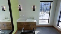 This single-floor space is designed with a clean, modern aesthetic that blends functionality with contemporary style. The focal point of the room is a sleek floating vanity crafted from warm wood cabinetry, topped with a crisp white solid-surface countertop and an integrated vessel sink with a polished chrome faucet. Ample drawer and cabinet storage beneath the vanity ensures a tidy, organized workspace.

Flanking the vanity are two striking wall-mounted sconce lights featuring a bold vertical design with warm and blue-toned illumination, casting a dramatic, layered glow against the soft green accent wall. The contrast between the warm wood tones, white surfaces, and green walls creates a refreshing and inviting atmosphere.

A large picture window positioned to the right floods the room with natural light and provides an open view of an outdoor seating area and surrounding urban architecture, enhancing the sense of space. The floor is finished in a neutral grey carpet, adding warmth underfoot. A professional reclining chair in the foreground suggests this space is purpose-built for personal care or grooming services, combining comfort with a polished, professional environment.