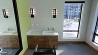 This single-floor space is designed with a clean, modern aesthetic that blends functionality with contemporary style. The focal point of the room is a sleek floating vanity crafted from warm wood cabinetry, topped with a crisp white solid-surface countertop and an integrated vessel sink with a polished chrome faucet. Ample drawer and cabinet storage beneath the vanity ensures a tidy, organized workspace.

Flanking the vanity are two striking wall-mounted sconce lights featuring a bold vertical design with warm and blue-toned illumination, casting a dramatic, layered glow against the soft green accent wall. The contrast between the warm wood tones, white surfaces, and green walls creates a refreshing and inviting atmosphere.

A large picture window positioned to the right floods the room with natural light and provides an open view of an outdoor seating area and surrounding urban architecture, enhancing the sense of space. The floor is finished in a neutral grey carpet, adding warmth underfoot. A professional reclining chair in the foreground suggests this space is purpose-built for personal care or grooming services, combining comfort with a polished, professional environment.