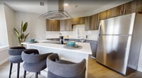 Located at 48–66 W Broad St in Rochester, NY, this 650-square-foot single-floor apartment offers a thoughtfully designed layout combining modern aesthetics with functional living. The open-concept kitchen and living area serves as the heart of the unit, showcasing a contemporary kitchen with wood-toned cabinetry, white subway tile backsplash, white quartz countertops, stainless steel appliances including a dishwasher and refrigerator, and a kitchen island for additional prep space. Light wood-style flooring runs throughout, creating a cohesive and airy feel. The living area is bright and inviting, featuring large windows that offer urban views of Rochester's downtown architecture, including a striking historic church tower. Natural light flows generously through the space, enhancing its open ambiance. The bedroom provides a comfortable retreat with continued light flooring and easy access to the bathroom. The bathroom is clean and modern, featuring a walk-in glass-enclosed shower with white subway tile, tile flooring, and a vanity. The overall design balances contemporary finishes with a warm, neutral palette, creating a stylish and comfortable urban living experience.