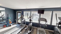 This single-floor fitness room is designed to provide a functional and focused workout environment. The space is defined by its bold navy blue walls, which create a clean, energetic atmosphere complemented by a textured ceiling and neutral-toned flooring. Multiple large windows fitted with horizontal blinds line the walls, allowing ample natural light to fill the room while offering views of the surrounding outdoor area. The generous influx of sunlight creates bright, dynamic patterns across the floor, enhancing the overall energy of the space. The room is equipped with a selection of cardio machines, including two treadmills and an elliptical trainer, arranged to make efficient use of the available floor space. A wall-mounted flat-screen television is positioned for easy viewing during workouts, adding an entertainment element to the exercise experience. Recessed ceiling lighting provides additional illumination, ensuring the space remains well-lit at all hours. A door provides direct access to the outdoors, further enhancing ventilation and connectivity to the exterior. Overall, this fitness room offers a practical, well-appointed setting suited for daily cardiovascular exercise.