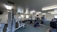 This single-floor fitness center is designed to accommodate a well-rounded workout experience. The space is outfitted with a diverse selection of exercise equipment, including treadmills, a stair climber, a stationary bike, and a multi-station strength training machine, along with a weight bench and additional free-standing equipment. The dark, textured flooring provides a stable and durable surface suited for high-traffic athletic use. Fluorescent lighting fixtures are evenly distributed across the textured ceiling, ensuring the room is brightly and uniformly lit throughout. Full-length mirrors line portions of the walls, visually expanding the space and allowing users to monitor their form during workouts. Windows along one wall allow for natural light to supplement the artificial lighting, offering views to the exterior. A wall-mounted television is also present, adding an entertainment element to the workout environment. The overall layout is open and practical, with equipment arranged to allow comfortable movement between stations. The space maintains a clean, functional aesthetic focused entirely on fitness utility.