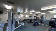 This single-floor fitness center is designed to accommodate a well-rounded workout experience. The space is outfitted with a diverse selection of exercise equipment, including treadmills, a stair climber, a stationary bike, and a multi-station strength training machine, along with a weight bench and additional free-standing equipment. The dark, textured flooring provides a stable and durable surface suited for high-traffic athletic use. Fluorescent lighting fixtures are evenly distributed across the textured ceiling, ensuring the room is brightly and uniformly lit throughout. Full-length mirrors line portions of the walls, visually expanding the space and allowing users to monitor their form during workouts. Windows along one wall allow for natural light to supplement the artificial lighting, offering views to the exterior. A wall-mounted television is also present, adding an entertainment element to the workout environment. The overall layout is open and practical, with equipment arranged to allow comfortable movement between stations. The space maintains a clean, functional aesthetic focused entirely on fitness utility.