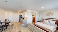 This efficiently designed one-bedroom unit offers a thoughtful layout that maximizes every square foot of its single-floor footprint. The open-concept living and kitchen area forms the heart of the space, where light wood-toned flooring runs seamlessly throughout, creating a bright and cohesive aesthetic. The kitchen is outfitted with modern two-toned cabinetry — white upper cabinets paired with gray lower units — complemented by white quartz countertops, a stainless steel sink, and a full suite of stainless steel appliances including a refrigerator, range, microwave, and dishwasher. Recessed lighting illuminates the kitchen, while a living area with a wall-mounted mini-split HVAC unit and ample natural light from a large window rounds out the shared space. A stylish sliding barn door with a frosted glass panel leads into the bedroom, which features a dedicated closet, a natural light-filled window with outdoor views, and clean white walls accented by decorative wall art. The bedroom's cozy scale and clean design make it a functional and restful retreat. Throughout the unit, the modern finishes, neutral palette, and smart use of space reflect a contemporary and practical design approach.