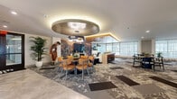 Located at 1724 Partnership Way in Houston, TX, this upscale social lounge offers an expansive, single-floor layout designed for both community gathering and individual productivity. The space is defined by its contemporary aesthetic, featuring a dynamic combination of polished stone tile flooring and richly patterned carpet tiles that visually delineate distinct activity zones throughout.

Upon entry, a sleek reception area welcomes visitors with warm artwork, a decorative console, and gleaming large-format tile floors. The main lounge area is anchored by a circular ceiling medallion with integrated ambient lighting and a statement pendant fixture, hovering above a central dining arrangement. Bold, large-scale murals adorn accent walls, adding artistic energy to the environment.

A fully equipped kitchen island with bar seating, pendant lighting, and wood-paneled ceiling accents serves as a central hub for social interaction. Multiple seating configurations — from collaborative workstations with white quartz surfaces to plush lounge groupings — provide versatile functionality throughout.

Floor-to-ceiling windows line one side of the space, flooding the interior with natural light and offering views of an outdoor terrace with additional seating. Warm gold-toned architectural niches and wood ceiling details add refined warmth, creating an inviting, resort-style atmosphere throughout.