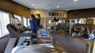 This fitness center offers a comprehensive workout environment designed to accommodate a variety of exercise routines. The space is laid out on a single level, with dedicated zones for both cardiovascular training and strength work. A row of treadmills lines one side of the room, providing ample space for cardio activity, while a selection of weight machines and strength-training equipment occupies the opposite area, visible through large mirrored walls that enhance the sense of depth and openness throughout the space. The mirrored walls serve both a practical and aesthetic purpose, allowing users to monitor their form while visually expanding the room. Recessed lighting in the ceiling, supplemented by ceiling fans, ensures the space remains well-lit and ventilated during workouts. The flooring features durable rubber matting, appropriate for high-traffic fitness use. Colorful abstract artwork adorns the upper walls, adding a touch of visual interest and energy to the environment. Natural light filters in through windows along one wall, contributing to a bright and welcoming atmosphere. Overall, the space is thoughtfully organized to support an efficient and comfortable fitness experience.