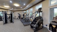 This fitness center offers a comprehensive workout environment designed to accommodate a wide range of exercise routines. The space is housed in a single-level room with tray ceilings accented by dark crown molding, warm neutral-toned walls, and durable patterned carpeted flooring throughout. Recessed lighting and flush-mount fixtures provide consistent illumination, while ceiling fans contribute to comfort during workouts.

Large windows positioned along the exterior walls invite natural light and offer views of the surrounding outdoor areas. The cardio section is well-stocked with treadmills, elliptical trainers, and a StairMaster step machine, all arranged to face the windows. Wall-mounted flat-screen televisions are positioned throughout the room for entertainment during exercise sessions.

The strength training area features a multi-station cable machine, a dedicated free weight section with a full dumbbell rack, adjustable benches, and a barbell rack with weight plates. A large wall mirror spans the free weight zone, visually expanding the space and aiding in form monitoring. Additional equipment includes medicine balls, stability balls, and a foam mat for functional training. The fitness center also provides direct access to an adjacent outdoor area, enhancing the overall experience.