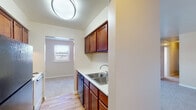 This single-floor residence offers a practical and comfortable layout across several well-defined living spaces. The galley-style kitchen is efficiently designed, featuring rich cherry-toned wood cabinetry, light countertops, a double stainless steel sink, and hardwood-style flooring. Appliances including a refrigerator and range are integrated seamlessly into the layout, with a doorway at the end providing easy flow to adjoining areas.

The bedrooms are generously sized, each finished with neutral-toned carpet that lends a warm, quiet atmosphere. Natural light filters in through multiple windows fitted with blinds, offering pleasant outdoor views of surrounding greenery. One bedroom features dual closets with panel doors, providing abundant storage. Ceiling fans are present in several rooms, supplementing the baseboard heating and wall-mounted air conditioning units for year-round comfort.

The bathroom is finished with sage green wall tiles, a pedestal sink with a mirrored medicine cabinet above, a full bathtub with shower, and wood-style vinyl flooring. The living area is open and bright, with neutral walls and carpet that create a clean, move-in ready canvas. Throughout the space, a consistent palette of warm neutrals and practical finishes defines an inviting and functional home.
